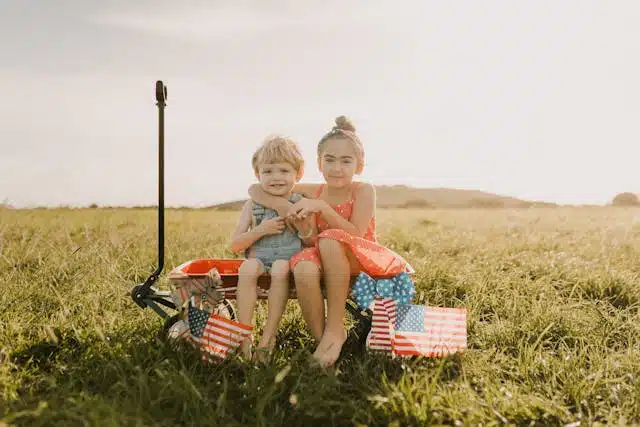 Deux jeunes enfants "host kids" souriants assis dans un champ avec des drapeaux américains, incarnant la vie quotidienne dans une famille d’accueil aux États-Unis